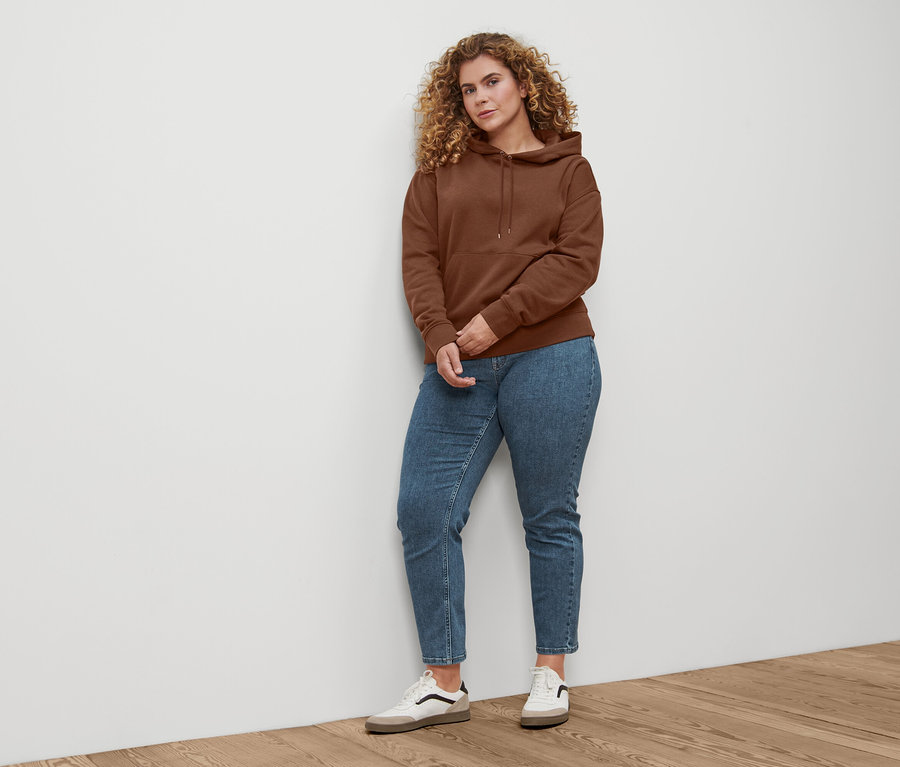 Une femme debout porte un jean droit en denim « Fit Juna » bleu moyen, des baskets en cuir et un sweat-shirt à capuche marron foncé.