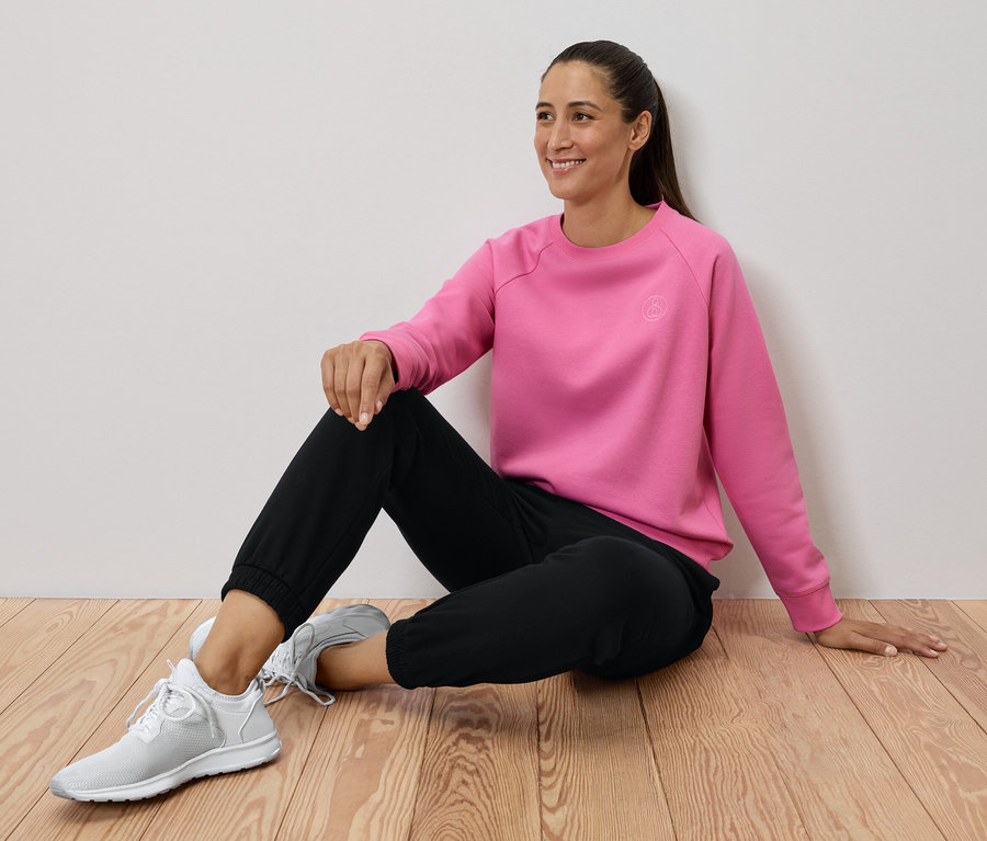 Femme assise sur un plancher en bois portant un sweat-shirt rose, un pantalon noir et des baskets blanches.
