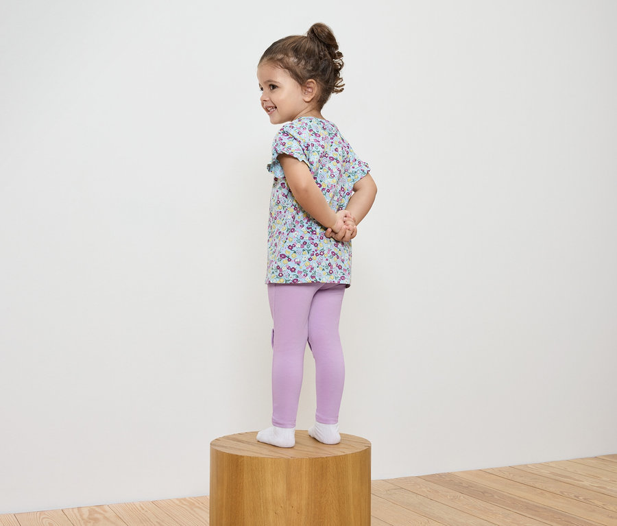 Une fille en blouse à fleurs et leggings violets se tient sur un socle en bois.