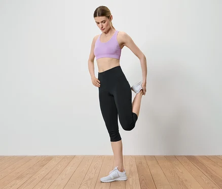 Une femme étire sa jambe en tenant sa cheville dans sa main. Elle porte un soutien-gorge de sport violet, un legging noir et des baskets blanches.