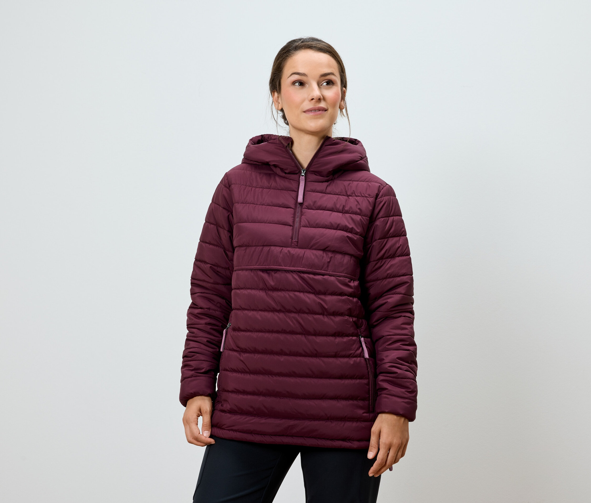 Une femme pose dans une veste matelassée bordeaux à capuche et un pantalon noir.