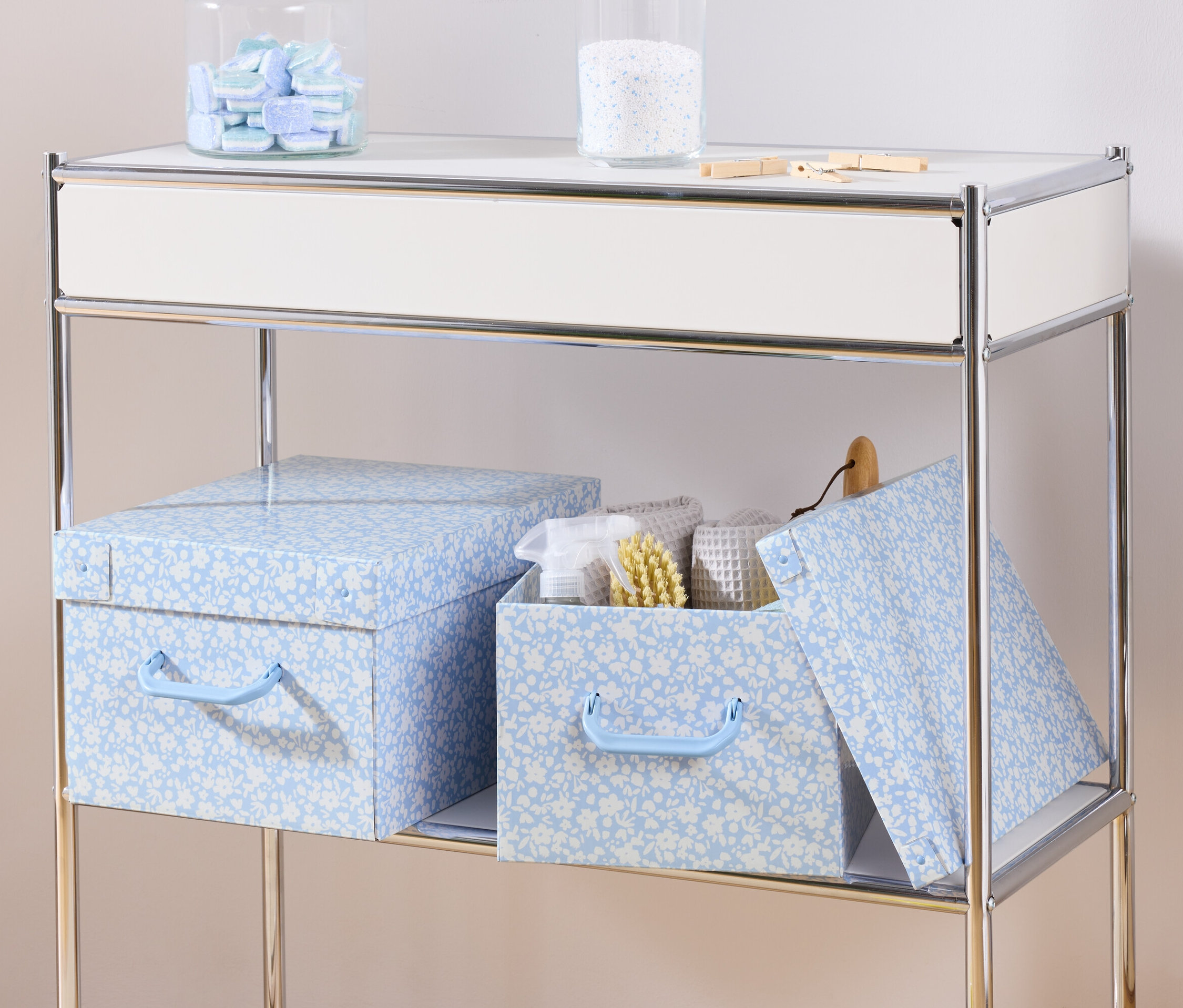 Sur une étagère, deux boîtes de rangement bleues avec couvercle et une boîte de rangement avec couvercle contenant des serviettes blanches et une brosse.