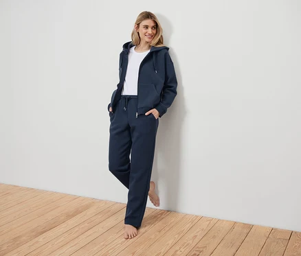 Femme en survêtement bleu marine appuyée contre un mur blanc.