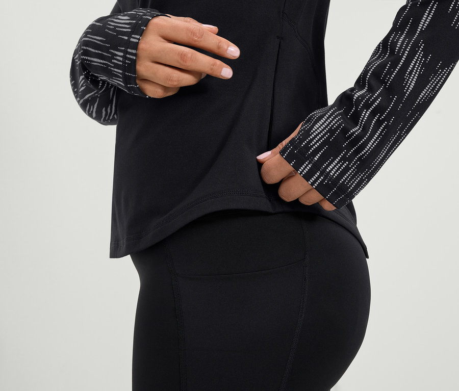 Une femme met la main dans la poche latérale d'un haut de sport noir à manches longues et d'un legging noir.