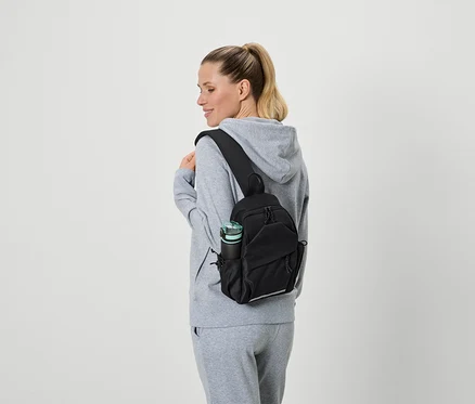 Femme portant un survêtement gris et un sac à bandoulière noir avec une bouteille d'eau dans la poche latérale.