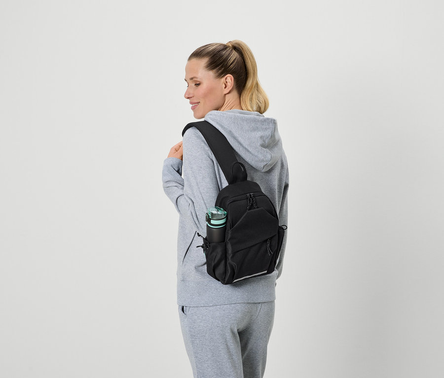 Femme portant un survêtement gris et un sac à bandoulière noir avec une bouteille d'eau dans la poche latérale.
