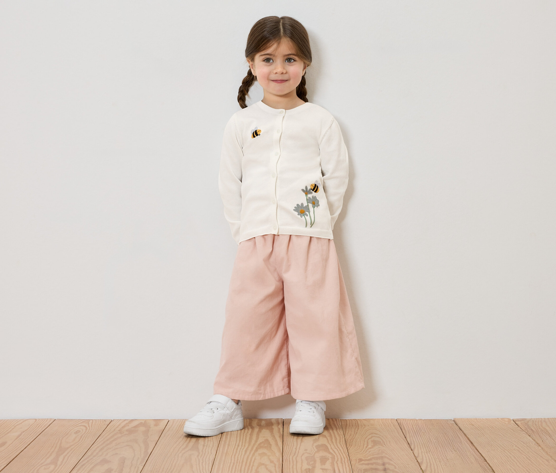 Une fillette avec des tresses porte un cardigan crème avec des broderies de fleurs et d'abeilles, un pantalon rose et des baskets blanches, devant un mur blanc.