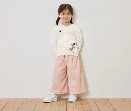 Une fillette avec des tresses porte un cardigan crème avec des broderies de fleurs et d'abeilles, un pantalon rose et des baskets blanches, devant un mur blanc.