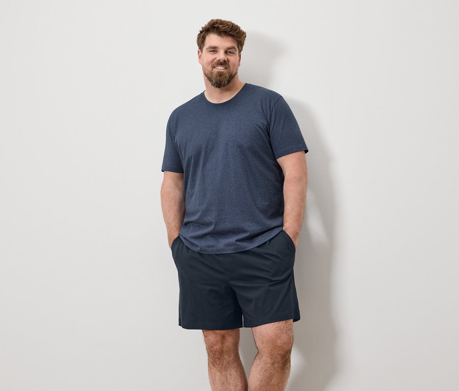 Un homme barbu se tient devant un mur blanc, les mains dans les poches, portant un t-shirt bleu et un short.