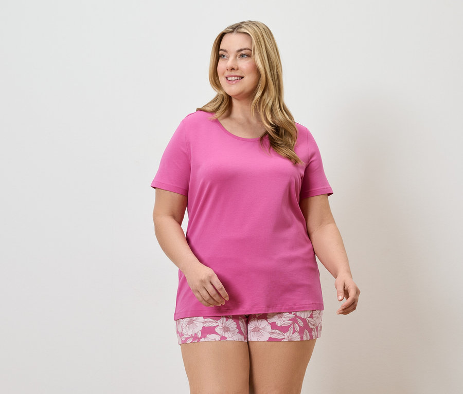Une femme blonde porte un t-shirt rose et un short à fleurs sur fond blanc.