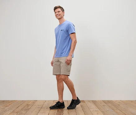 Un homme marche sur un plancher en bois, vêtu d'un t-shirt bleu, d'un short beige et de baskets noires.