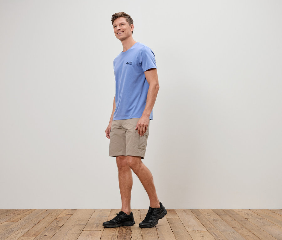 Un homme marche sur un plancher en bois, vêtu d'un t-shirt bleu, d'un short beige et de baskets noires.