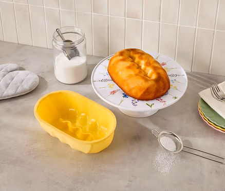 Sur un comptoir de cuisine, on trouve un kouglof sur un présentoir à fleurs, un moule à kouglof jaune, un pot de sucre, un tamis et des maniques.