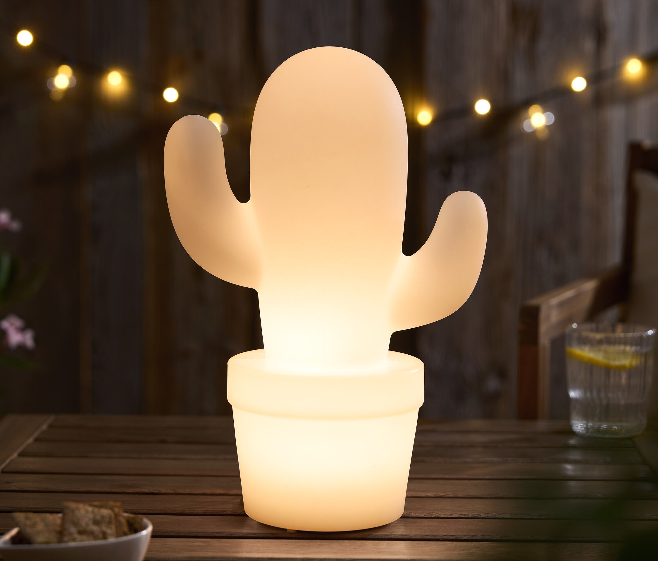 Cactus lumineux dans un pot sur une table en bois, avec une guirlande lumineuse en arrière-plan.