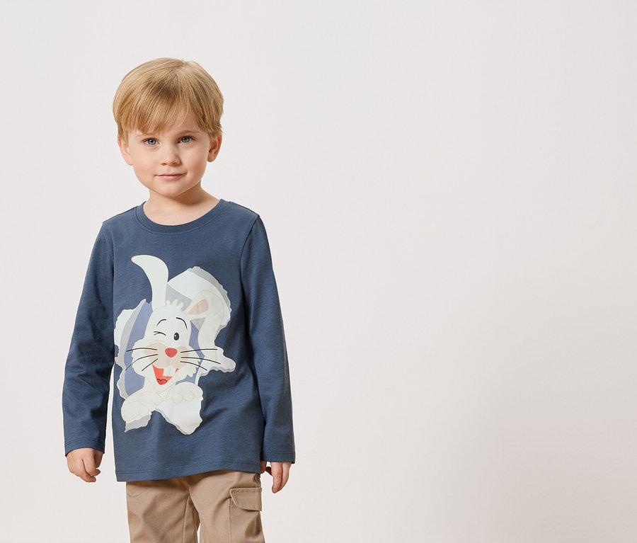 Un petit garçon porte un T-shirt bleu à manches longues avec un imprimé de lapin blanc et un pantalon beige.