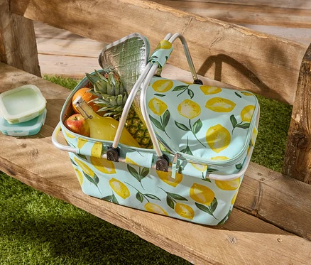 Un sac de pique-nique à motif citron est posé sur un banc en bois, rempli de fruits, d'une bouteille et de deux boîtes de conservation.