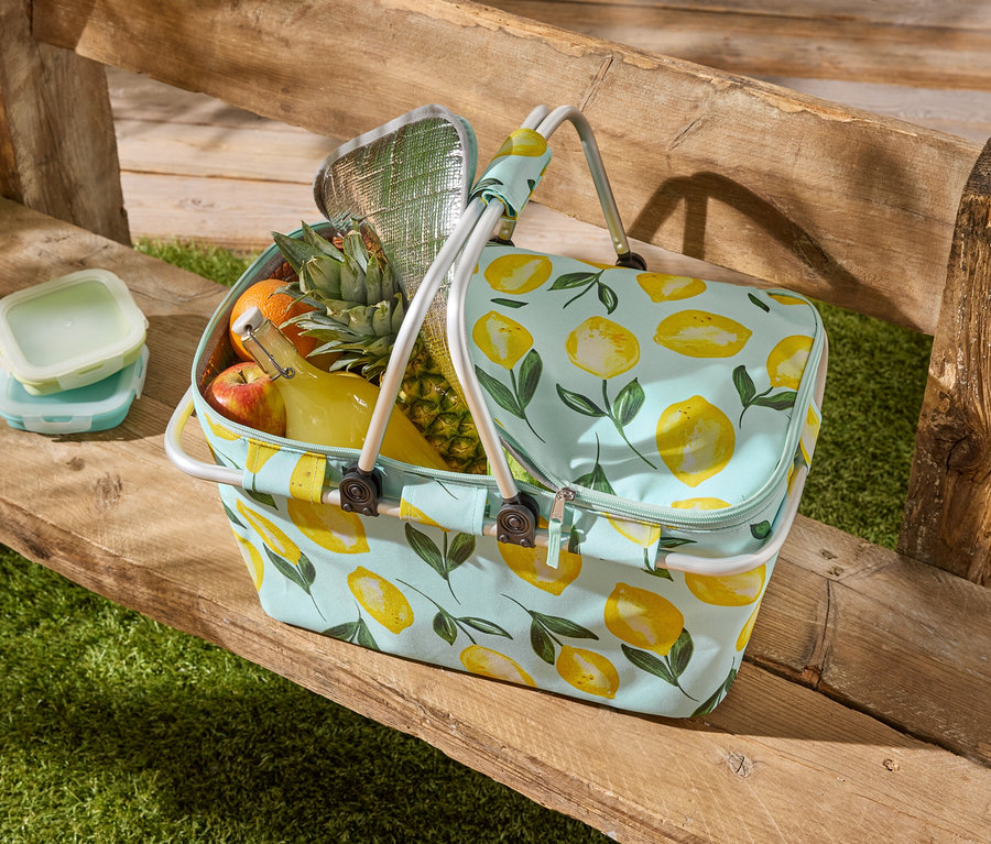 Un sac de pique-nique à motif citron est posé sur un banc en bois, rempli de fruits, d'une bouteille et de deux boîtes de conservation.