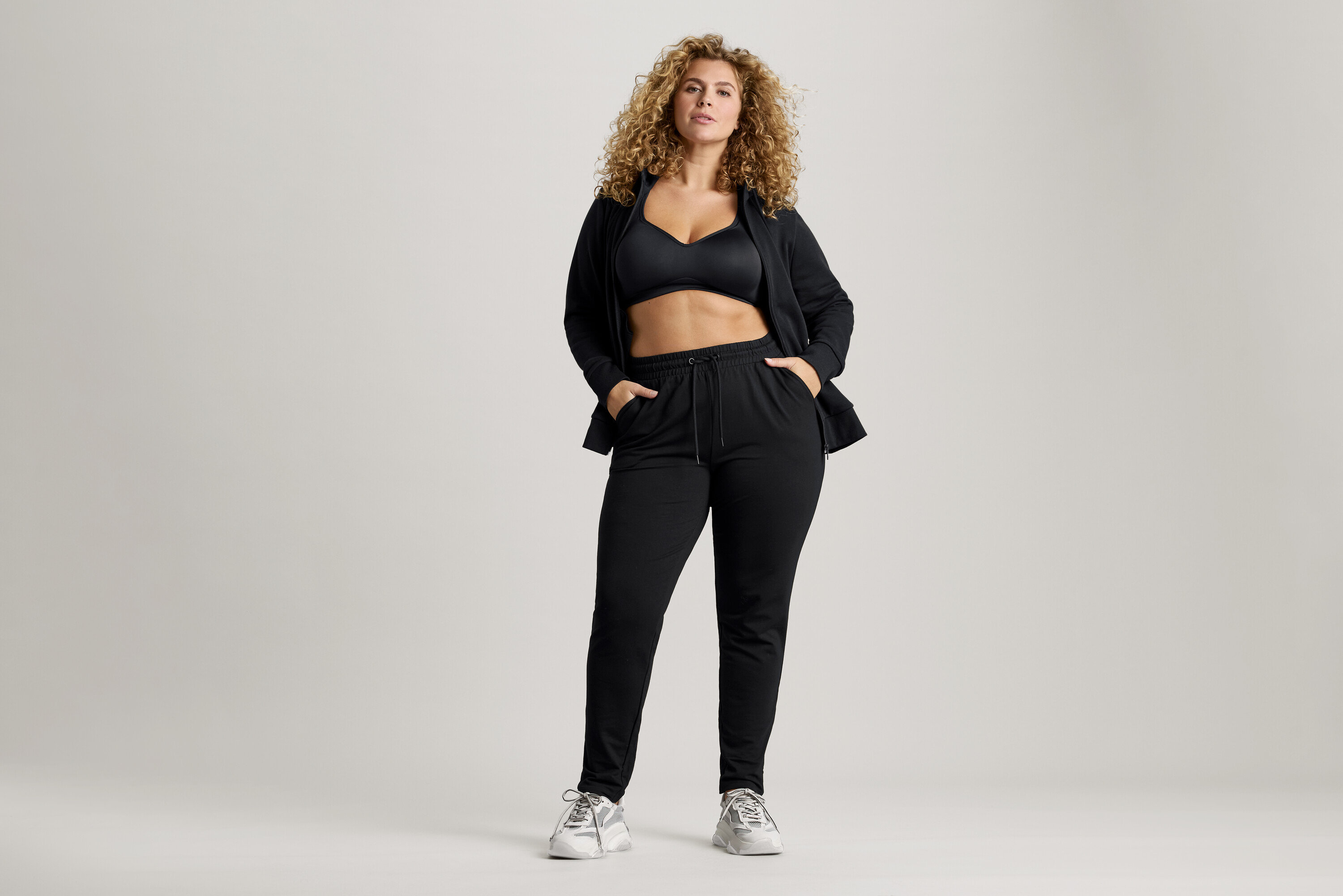 Femme aux cheveux bouclés portant un soutien-gorge de sport noir, une veste et un pantalon de survêtement avec les mains dans les poches.