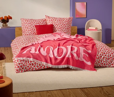 Sur un lit à motif léopard rouge et blanc se trouve une couverture rouge avec le mot "AMORE" en grandes lettres blanches.