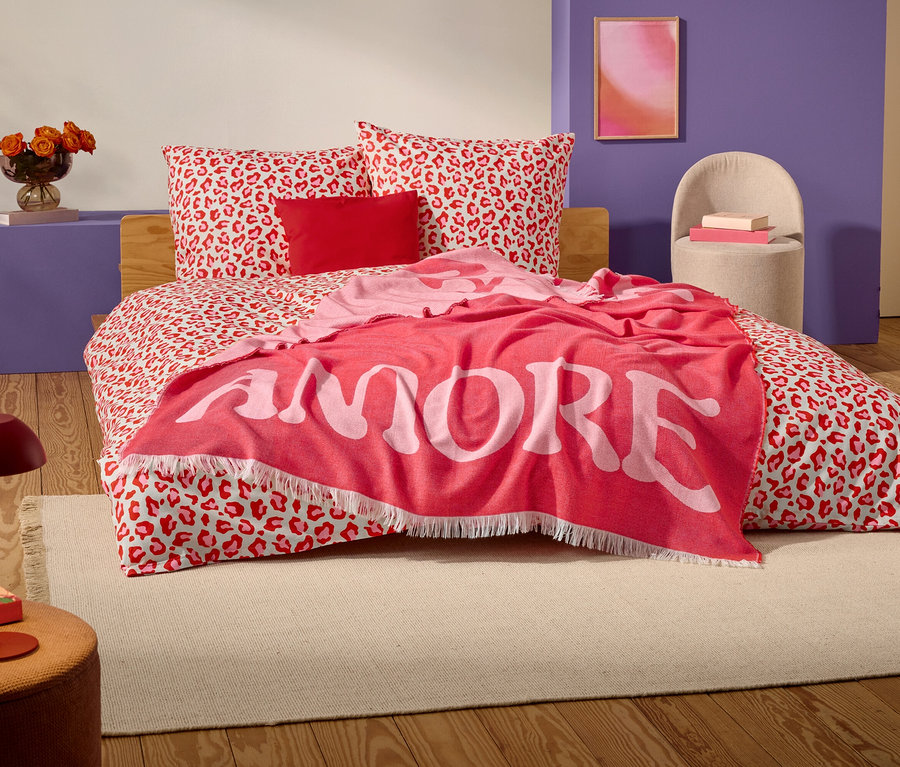 Sur un lit à motif léopard rouge et blanc se trouve une couverture rouge avec le mot "AMORE" en grandes lettres blanches.