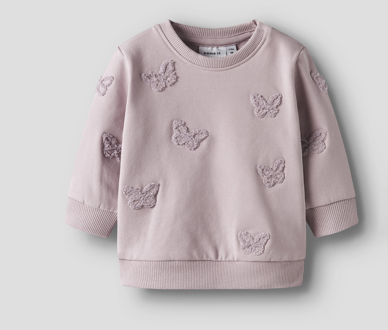 Un sweat-shirt pour bébé NAME IT lilas avec des papillons cousus est posé sur un fond neutre.