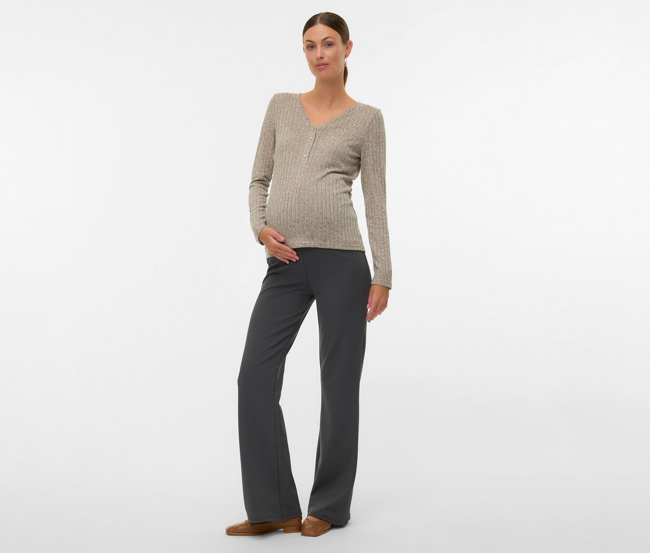 Femme avec un chignon portant un T-shirt MAMALICIOUS «
MLPAMMIE LIA », un pantalon gris, des chaussures marron, main sur le ventre.