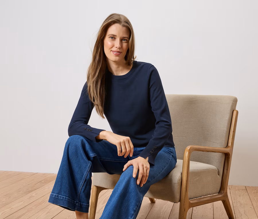 Une femme aux cheveux longs est assise sur une chaise, portant un T-shirt à manches longues bleu marine et un jean ample.