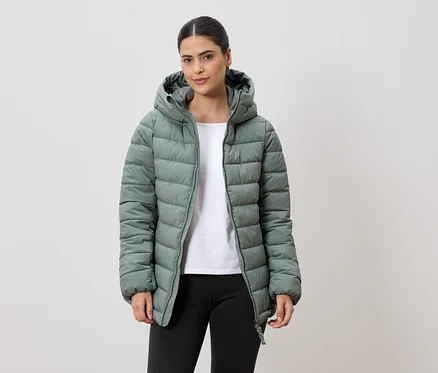 Une femme avec une queue de cheval porte une veste matelassée vert clair à capuche, un t-shirt blanc et un legging noir sur fond blanc.