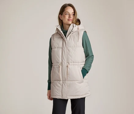 Femme aux cheveux châtains clairs, regardant vers la droite. Elle porte une longue doudoune sans manches beige clair à capuche, un haut à manches longues rayé vert et blanc et un pantalon foncé. Ses mains sont dans les poches du gilet.
