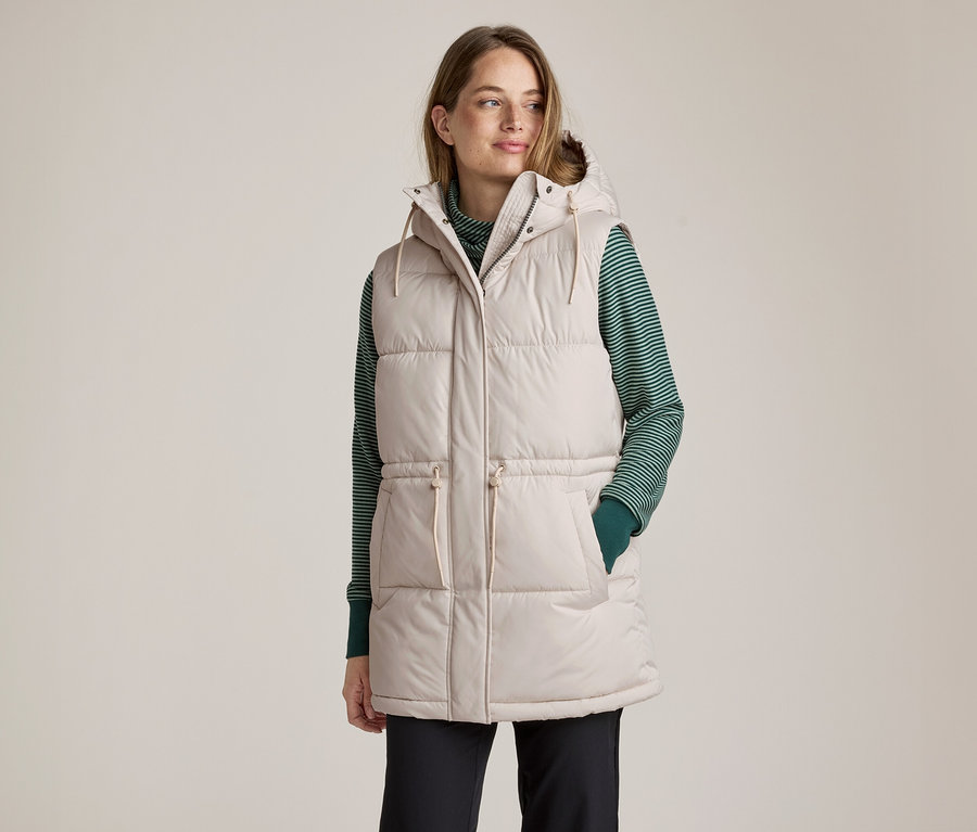 Femme aux cheveux châtains clairs, regardant vers la droite. Elle porte une longue doudoune sans manches beige clair à capuche, un haut à manches longues rayé vert et blanc et un pantalon foncé. Ses mains sont dans les poches du gilet.