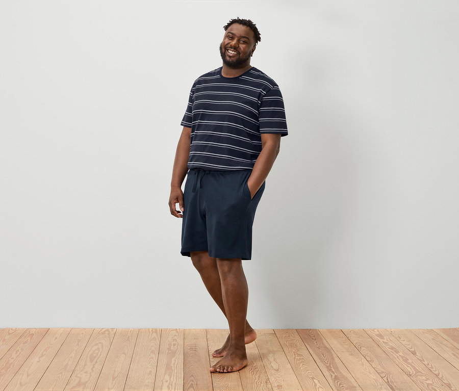 Un homme souriant se tient pieds nus devant un mur blanc, vêtu d'un t-shirt rayé et d'un short bleu marine.