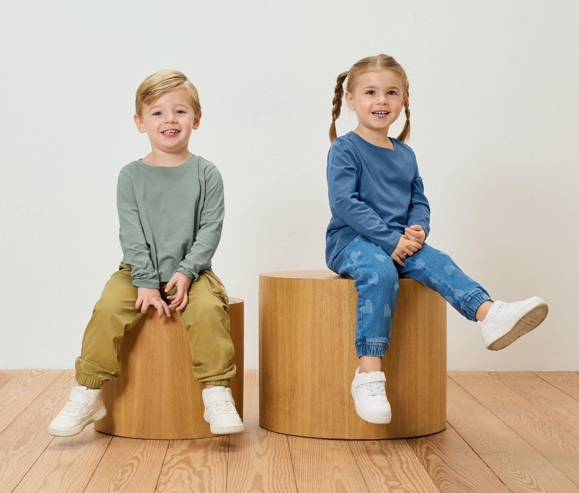 Un garçon avec un t-shirt à manches longues vert et un pantalon kaki est assis à côté d'une fille avec un t-shirt à manches longues bleu et un jean à motifs de cœur sur des tabourets en bois.