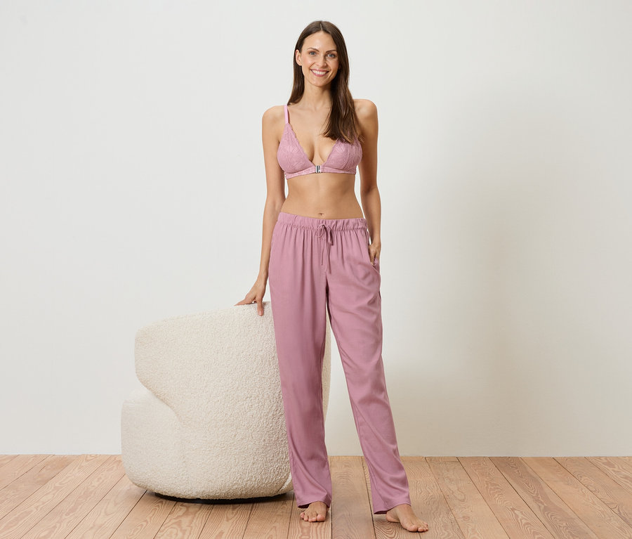Une femme se tient pieds nus à côté d'un fauteuil blanc, vêtue d'un soutien-gorge en dentelle rose et d'un pantalon rose ample.