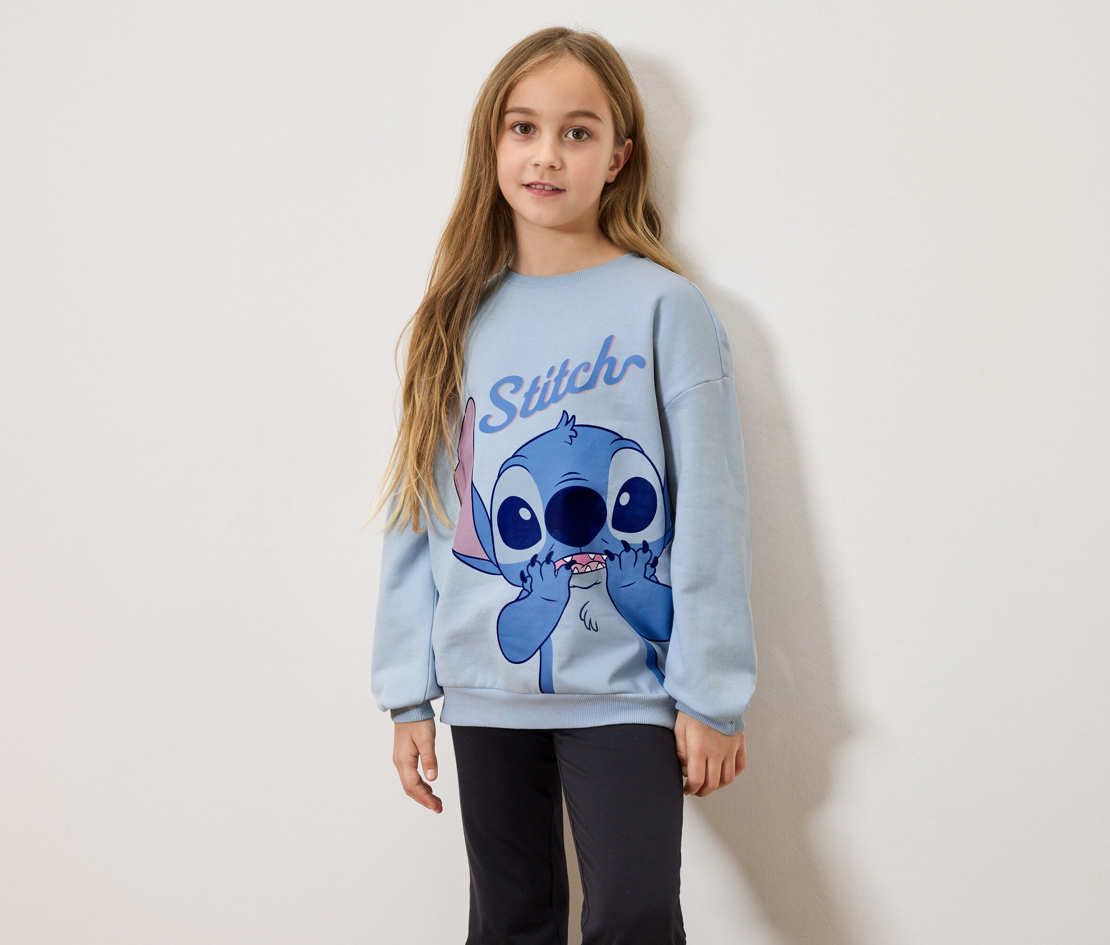 Une fille aux longs cheveux blonds porte un sweat-shirt bleu clair avec un imprimé Stitch et un legging noir.