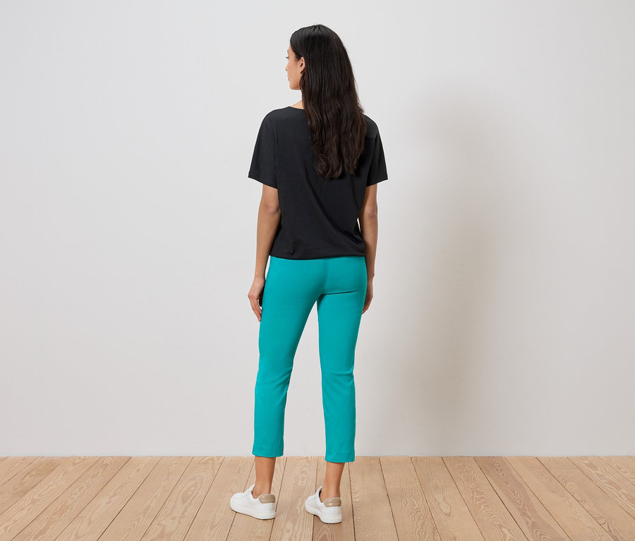 Vue de dos d'une femme aux cheveux noirs mi-longs portant un t-shirt noir, un pantalon turquoise et des baskets blanches.