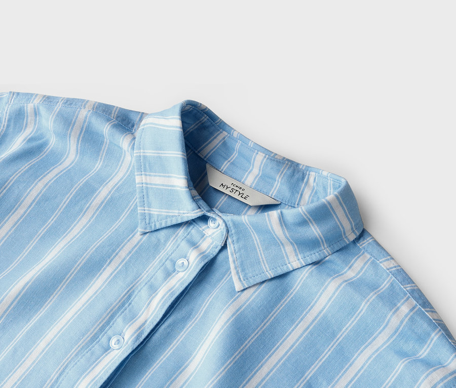 Gros plan d'une chemise rayée bleu clair et blanc avec un col et des boutons. Une étiquette indiquant « TCHIBO MY STYLE » est visible.
