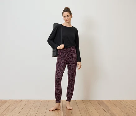 Une femme portant un chignon porte un haut noir, un pantalon à motifs et un tapis de yoga sur l'épaule, pieds nus sur un plancher en bois devant un mur blanc.