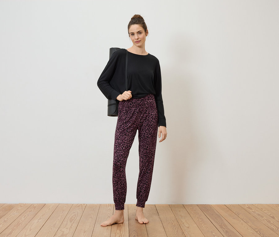 Une femme portant un chignon porte un haut noir, un pantalon à motifs et un tapis de yoga sur l'épaule, pieds nus sur un plancher en bois devant un mur blanc.