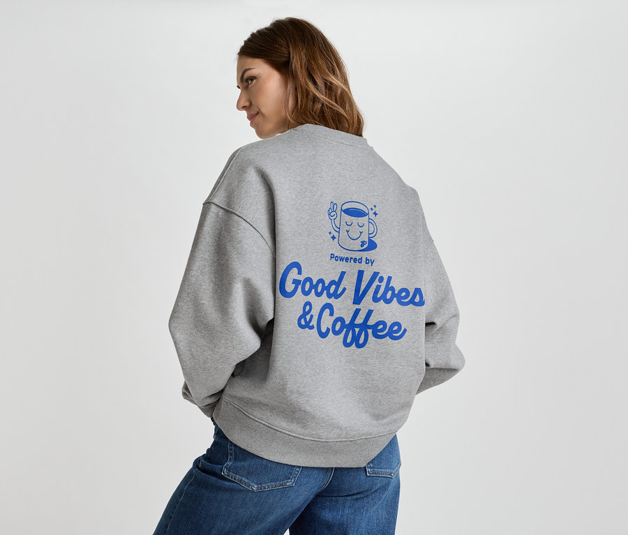 Femme portant un sweat-shirt gris avec l'inscription bleue "Good Vibes & Coffee" dans le dos, avec un jean.
