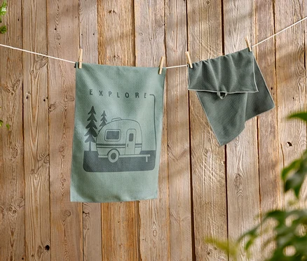 Un torchon de cuisine vert avec un motif de caravane est suspendu à côté d'une serviette texturée sur une corde à linge devant un mur en bois.
