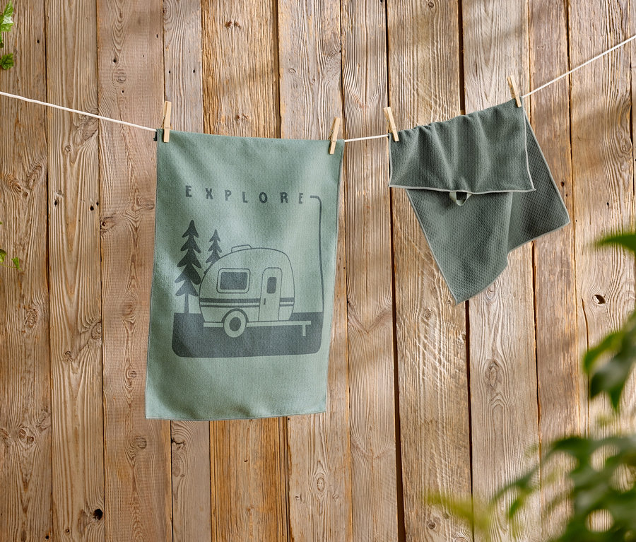 Un torchon de cuisine vert avec un motif de caravane est suspendu à côté d'une serviette texturée sur une corde à linge devant un mur en bois.