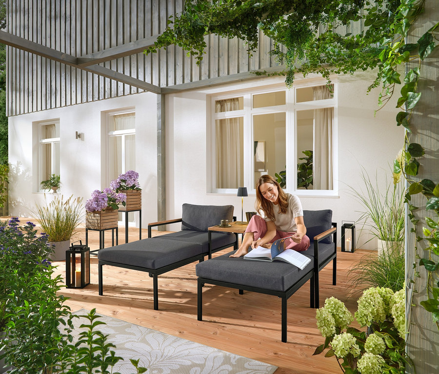 Une femme est assise sur une chaise longue de jardin grise et lit un livre. À l'arrière-plan, on peut voir deux colonnes à plantes « Elin ».