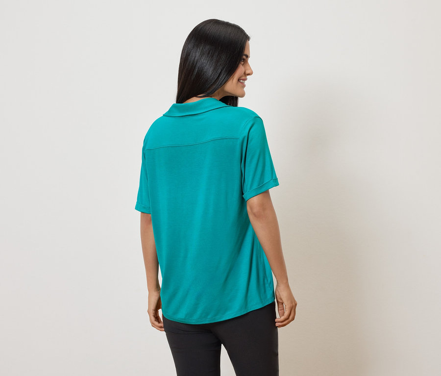Vue arrière d'une femme aux cheveux foncés portant un polo vert et un pantalon noir sur un fond crème.
