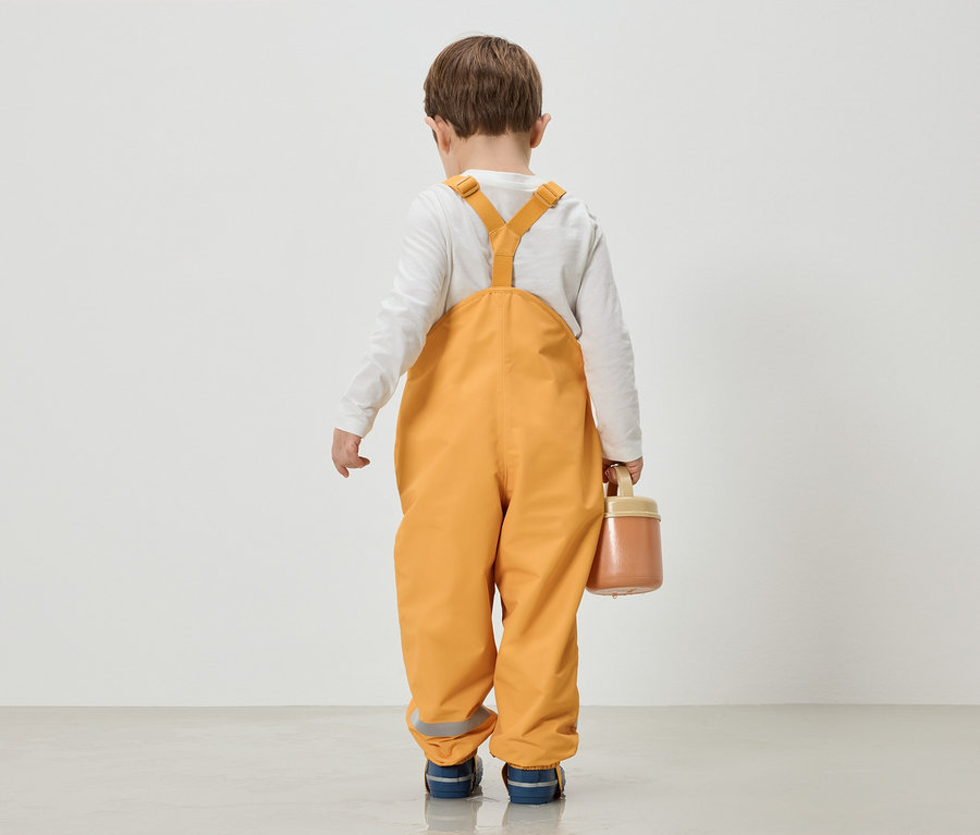 Un enfant en salopette jaune et chemise à manches longues blanche se tient dos à la caméra et tient une boîte.