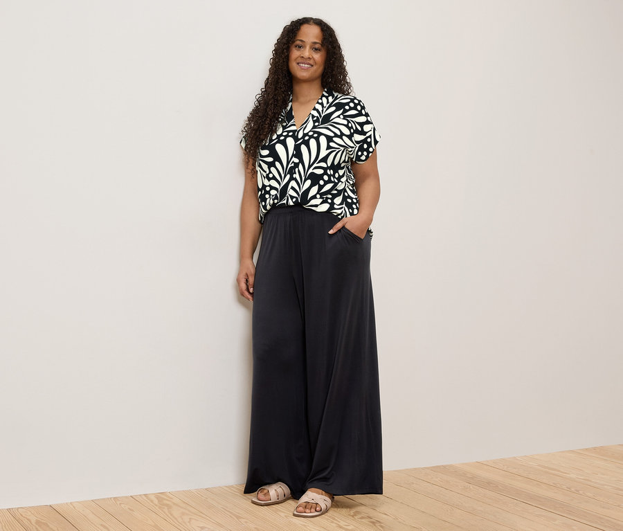 Une femme aux cheveux bouclés, portant un haut à motifs noirs et blancs et un pantalon ample foncé, se tient devant un mur.