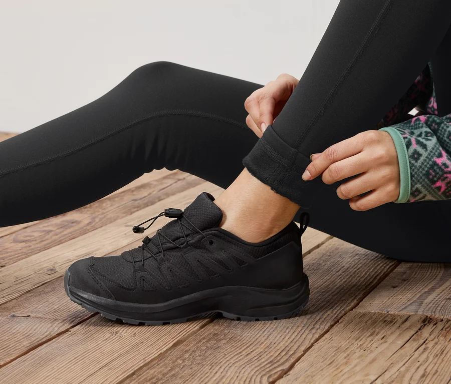 Une personne est assise sur un plancher en bois et remonte son legging noir, à côté d'une chaussure à lacets noire.