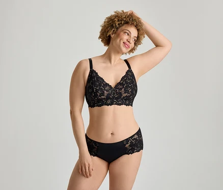 Une femme souriante aux cheveux bruns bouclés porte un soutien-gorge en dentelle noire et une culotte en dentelle noire assortie, posant une main derrière la tête.