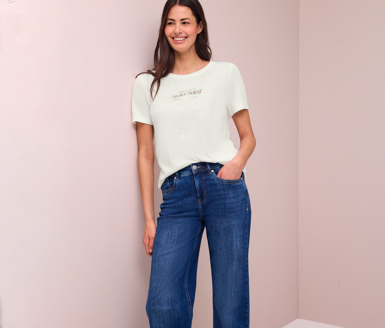 Une femme souriante, vêtue d'un jean et d'un t-shirt imprimé "Atelier Soleil", est appuyée contre un mur.