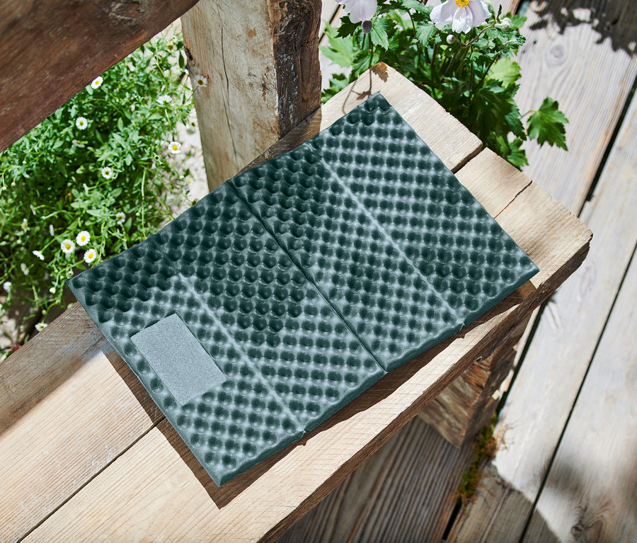 Un tapis de sol pliable vert foncé avec un motif à picots et une fermeture velcro grise est posé sur un banc en bois, entouré de plantes.