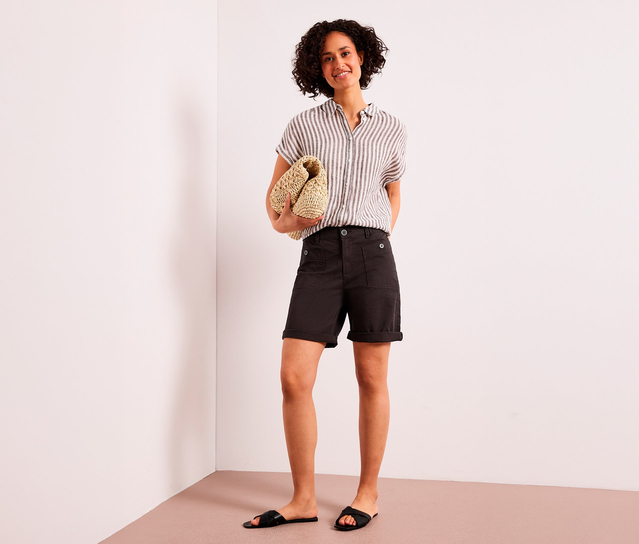 Une femme souriante aux cheveux bouclés se tient debout, tenant une pochette tressée. Elle porte une chemise rayée, un bermuda Street One « Yulius » marron et des sandales noires.